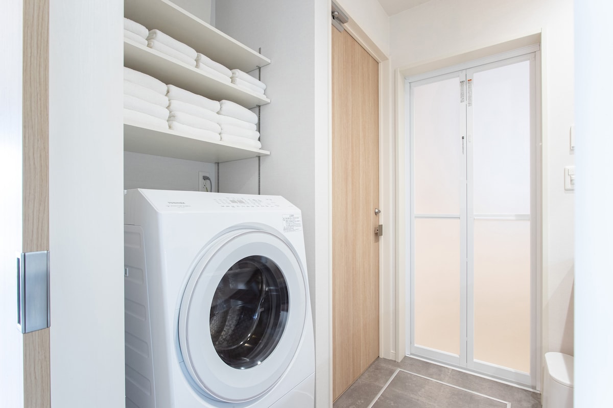A laundry area features a modern washing machine, complemented by neatly stacked towels on shelves above. A sliding door reveals additional space, enhancing functionality in the layout. The room's design incorporates neutral tones and ample natural light from the window.