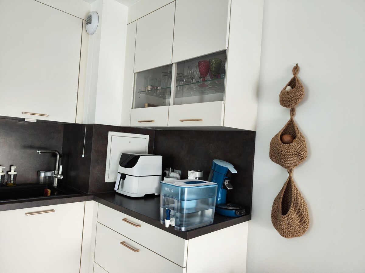 A modern kitchen features sleek white cabinetry with gold accents. A dark countertop is equipped with a water dispenser and kitchen appliances, including a coffee maker. Woven storage baskets hang from the wall, providing a decorative and functional element. Natural light brightens the space.