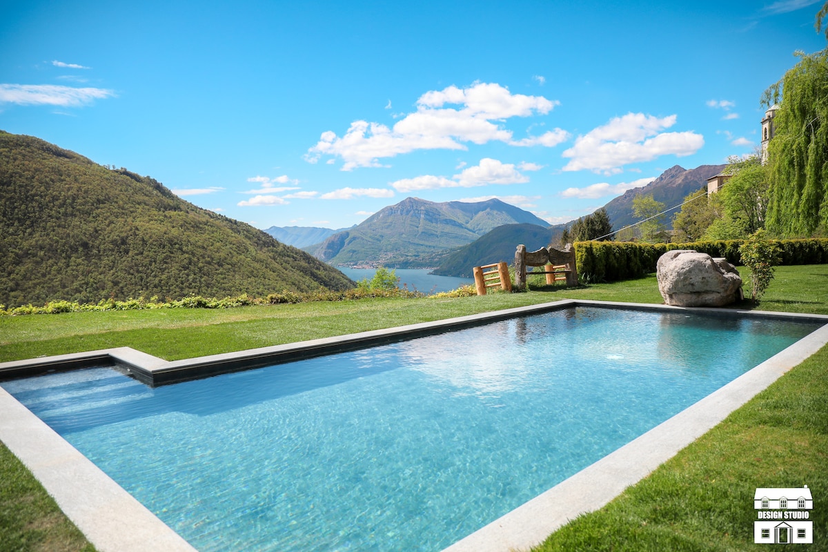 A tranquil outdoor pool is framed by lush greenery, offering panoramic views of the surrounding mountains and the lake. The clear blue water reflects the sky, while a large, smooth rock adds a natural touch to the landscape.