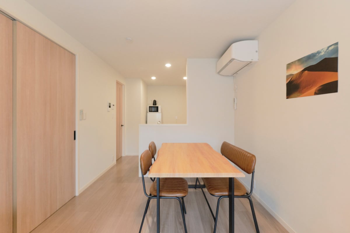 A dining area is presented with a wooden table accompanied by four brown chairs. Soft lighting creates a bright space with a view of an air conditioning unit mounted on the wall. A small kitchen area is visible in the background, showcasing modern appliances.