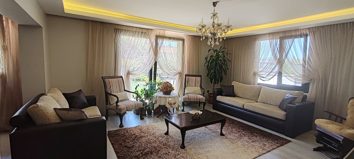A spacious living room features soft, neutral-toned furnishings arranged around a decorative coffee table. Large windows are adorned with sheer curtains, allowing natural light to filter in. A lush indoor plant adds a touch of greenery, and a chandelier provides an elegant overhead accent.