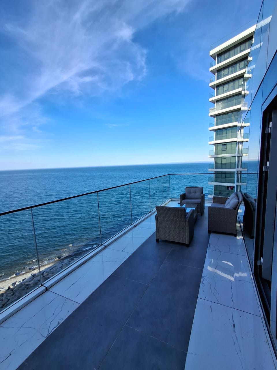 A spacious balcony overlooks the shimmering expanse of the ocean. Comfortable seating is provided by wicker chairs and a sofa, contrasting with the modern metal and glass railing. A clear blue sky stretches above, enhancing the serene and inviting outdoor space.