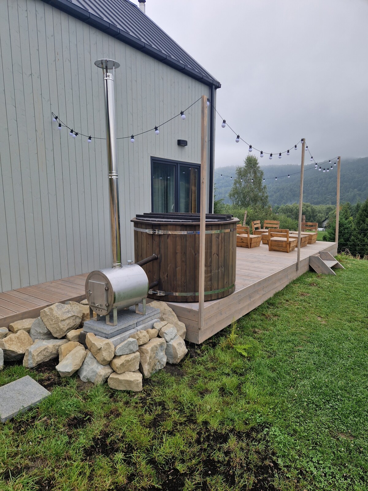 A wooden hot tub is positioned on a raised wooden deck, complemented by a stone base. The surrounding area features a set of wooden chairs arranged for relaxation, with green grass and a scenic view of the distant hills and cloudy sky.