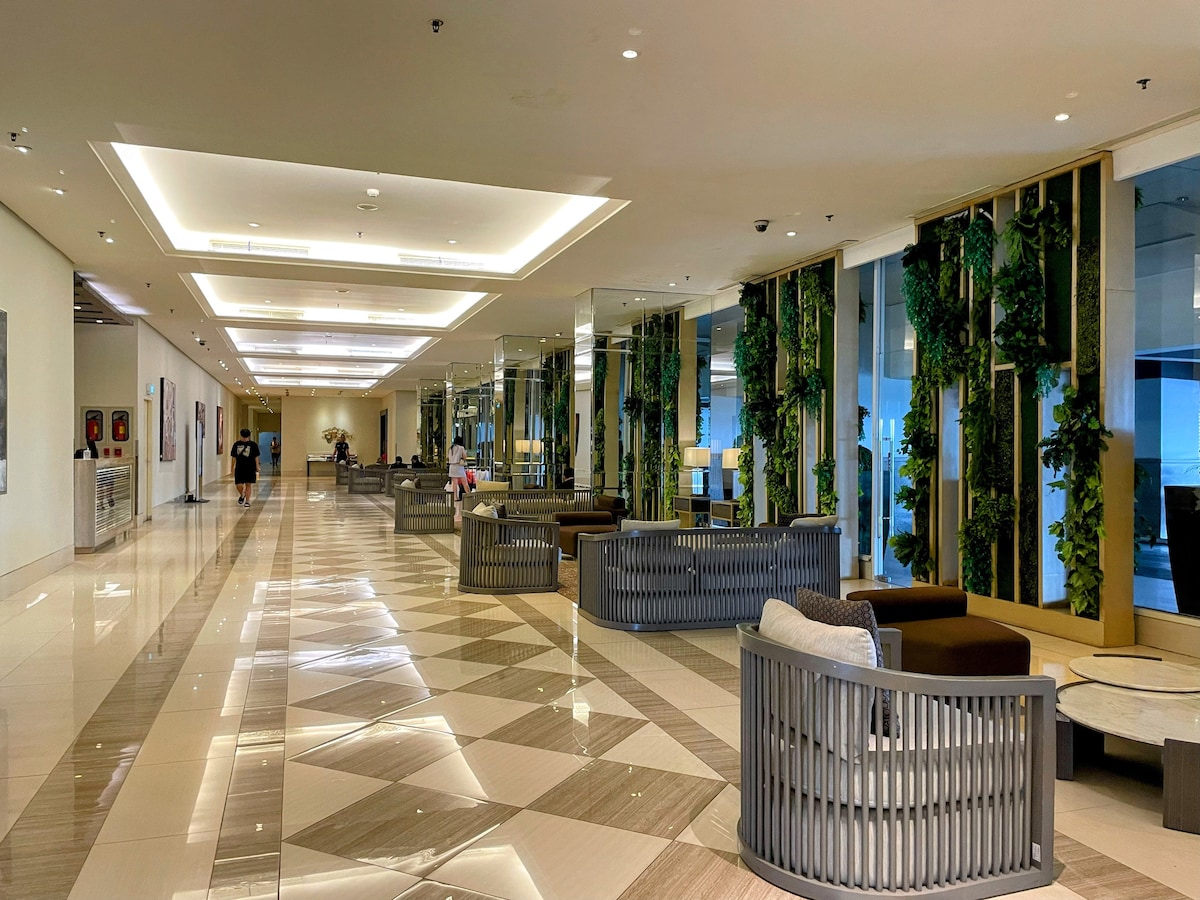A spacious lobby area features modern furnishings and expansive glass walls. Lush greenery is incorporated into the design, creating a fresh atmosphere. Soft seating arrangements offer comfort, and ambient lighting enhances the welcoming space, inviting guests for relaxation or socialization.