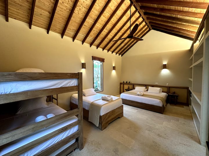 Habitación rústica de estilo caribeño con detalles naturales, muebles de madera y tonos cálidos. Incluye una cama tamaño king y cuatro camas individuales, perfectas para familias o grupos que buscan comodidad y tranquilidad en un ambiente tropical acogedor.
