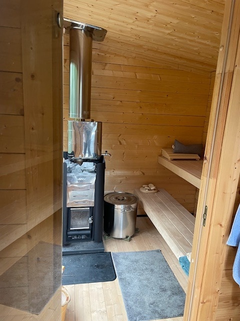 A serene interior of a sauna is presented, showcasing smooth wooden walls and benches. A metal stove is located prominently, with a small seating area to the side. Soft towels are neatly arranged on the benches, and a rug is placed at the entrance.