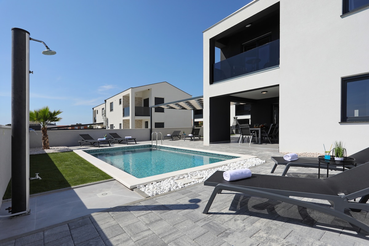 A modern outdoor area features a private pool surrounded by a tiled deck. Lounge chairs are arranged nearby, and a small patch of grass adds greenery. The villa's sleek architecture and large windows can be seen in the background beneath a clear blue sky.