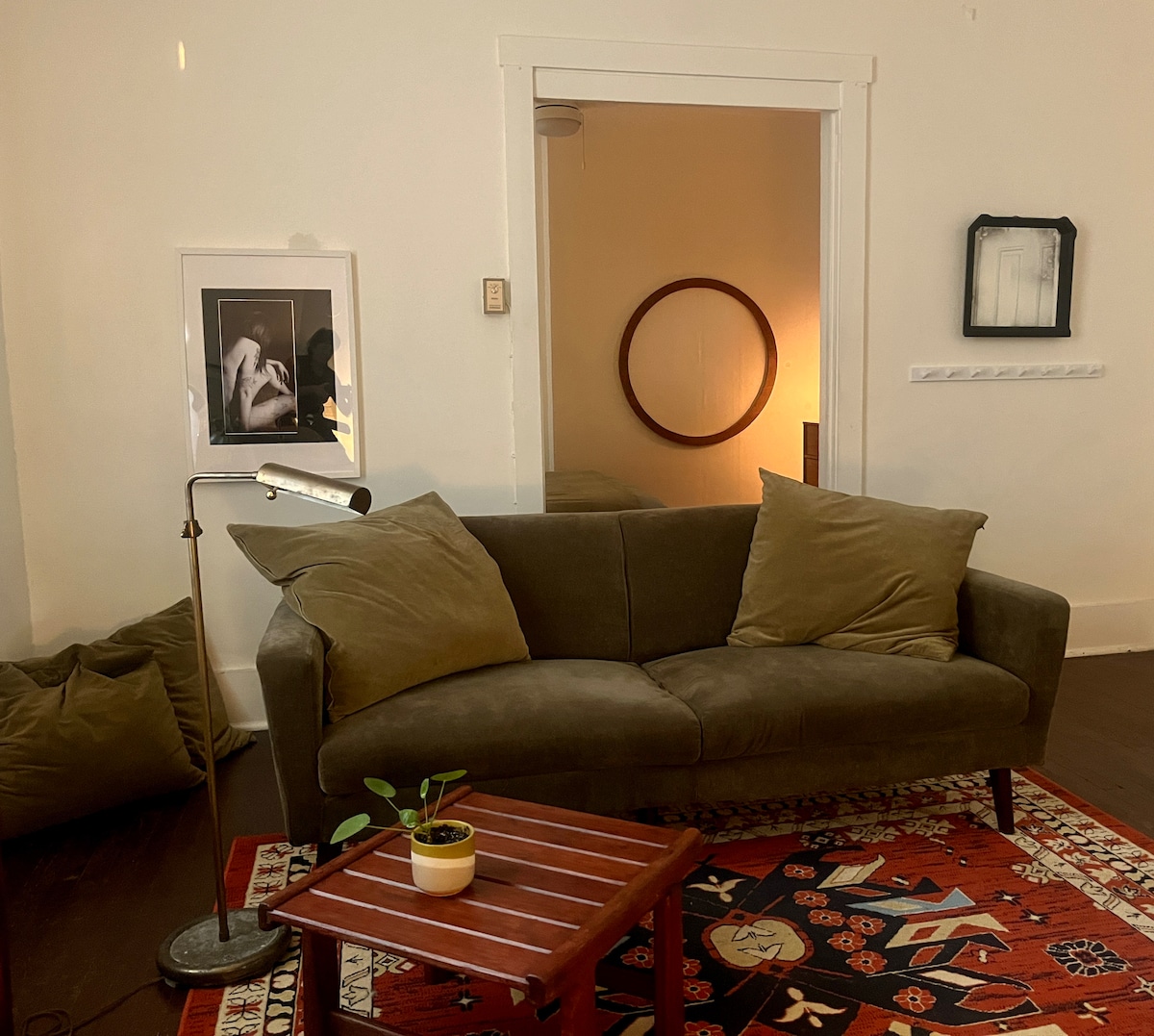 A minimalist living area features a cozy sofa with plush green cushions and a small wooden coffee table adorned with a potted plant. A warm glow emanates from a nearby lamp, while artwork and a circular mirror enhance the space's inviting atmosphere.