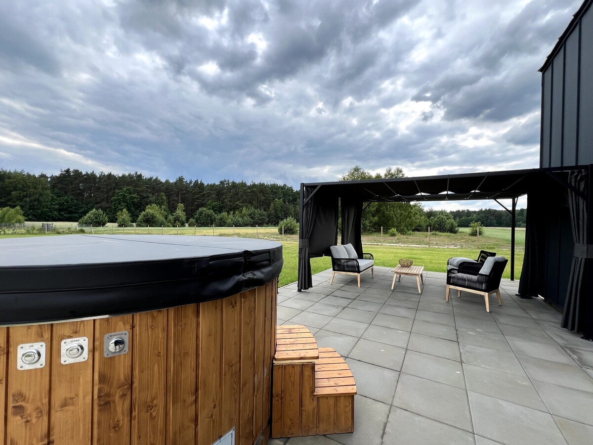 A modern outdoor area is showcased, featuring a wooden hot tub and a covered seating space. Two black chairs and a small table are placed on the patio, with an expansive grassy field and forest visible in the background under a cloudy sky.