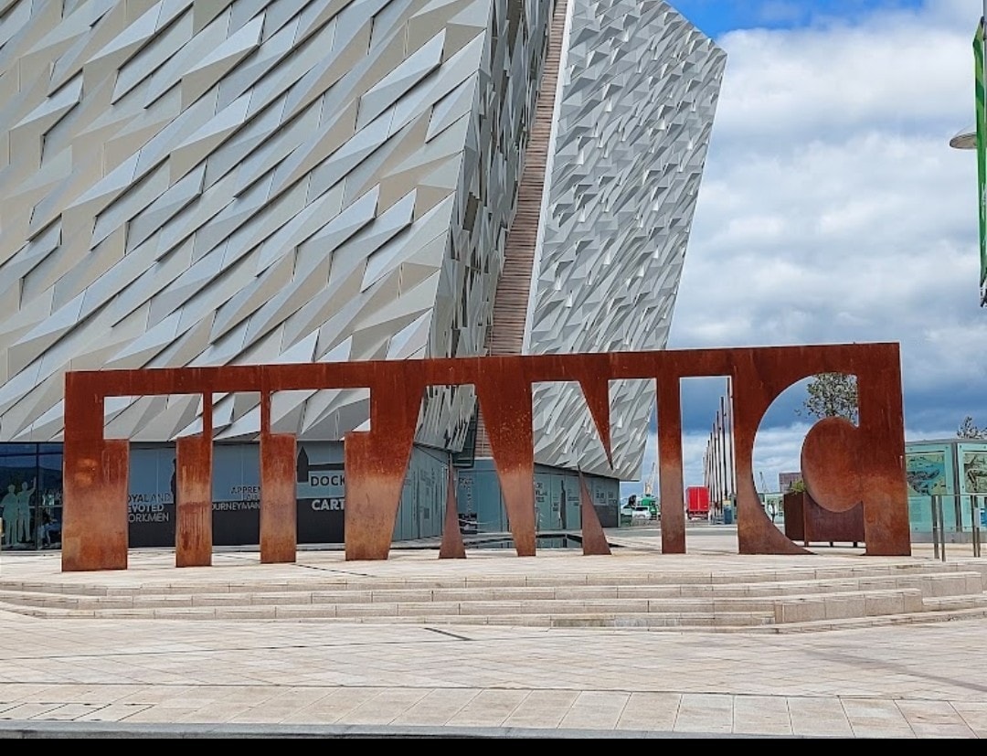 A large rust-colored metal sculpture displays the word 'TITANIC,' positioned prominently in front of a modern architectural structure with textured white exterior walls. The scene is set against a bright sky with scattered clouds.