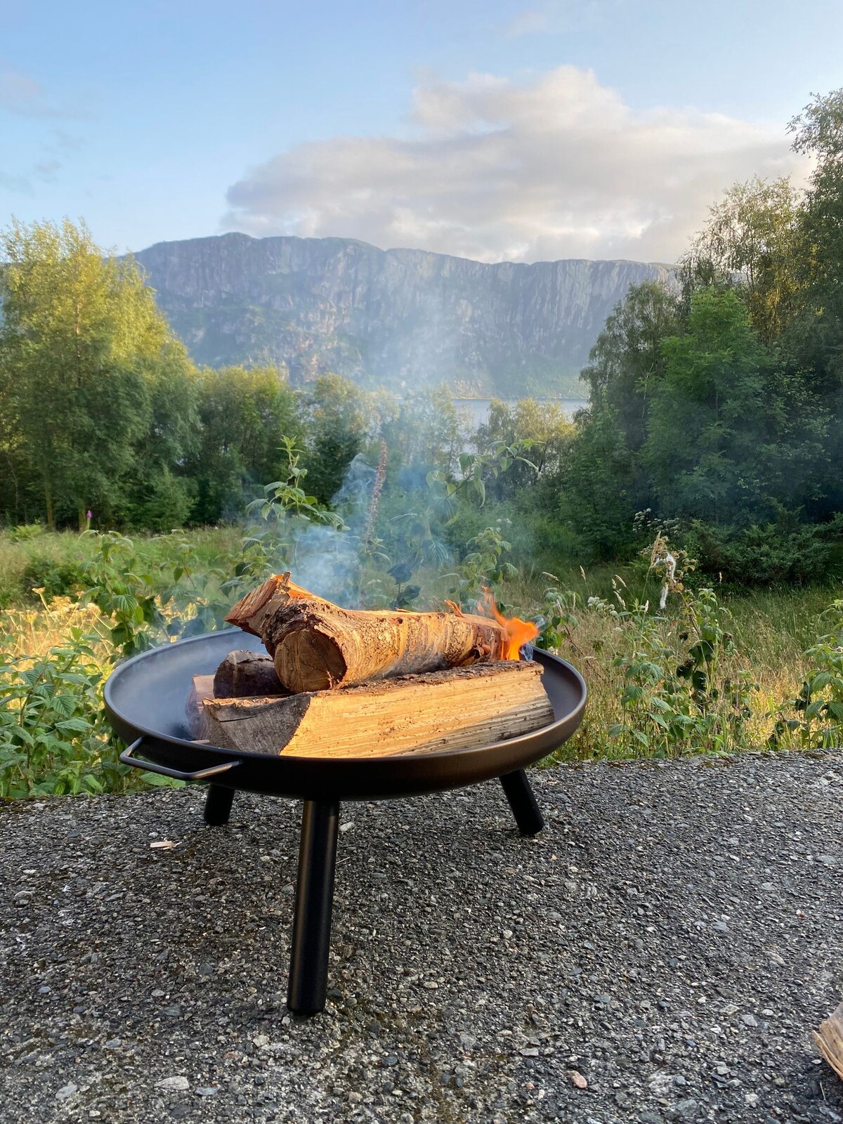 A fire pit sits on a stone surface, containing neatly stacked logs and gentle flames. The backdrop features rolling green hills and mountains under a partially cloudy sky, with the reflection of the landscape visible in a serene body of water.