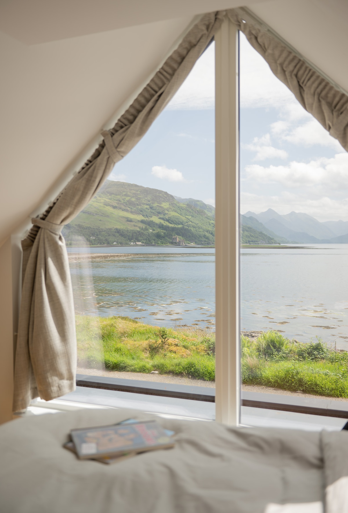 A large window with soft curtains frames scenic views of the calm waters of Loch Alsh and the lush green hills beyond. The natural light brightens a cozy corner featuring a spacious bed, creating a serene atmosphere.