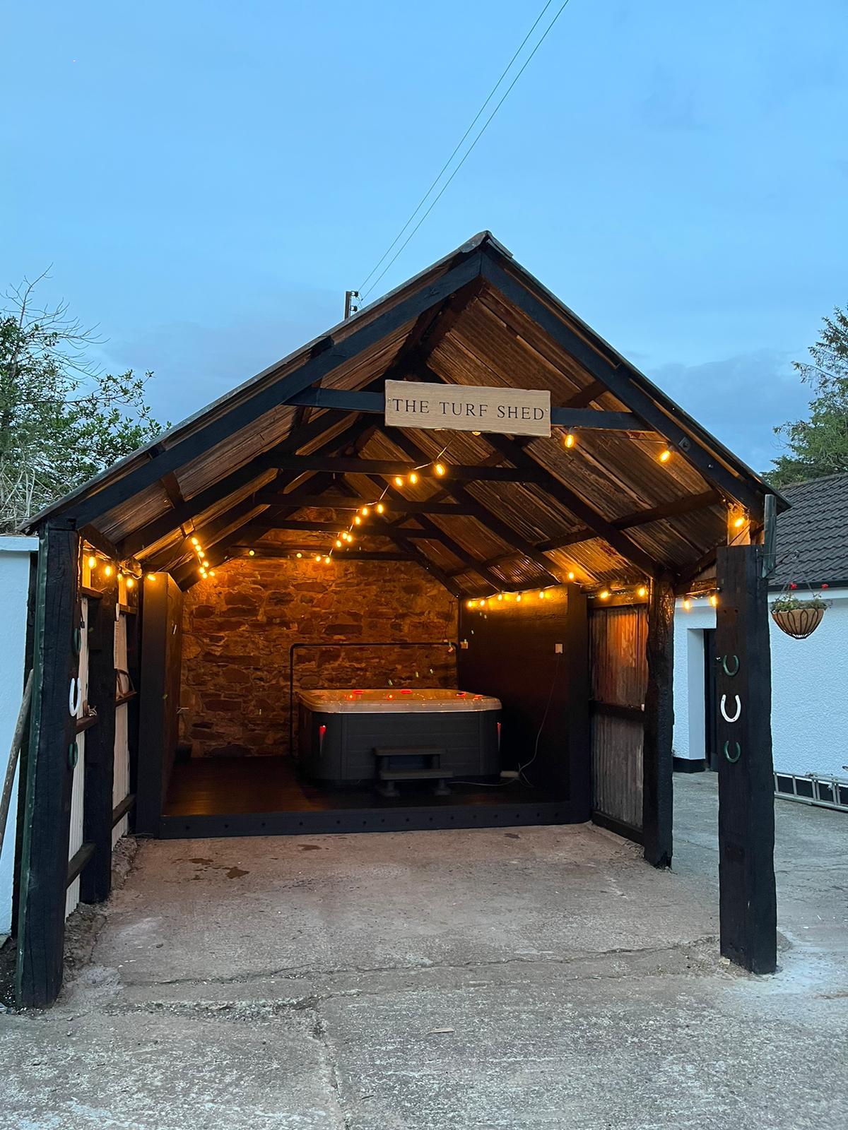 The image displays a rustic shed with a peaked roof and wooden beams. Soft string lights provide ambient lighting, illuminating a hot tub positioned inside. The stone wall adds character, contributing to a cozy and relaxing atmosphere suitable for unwinding.