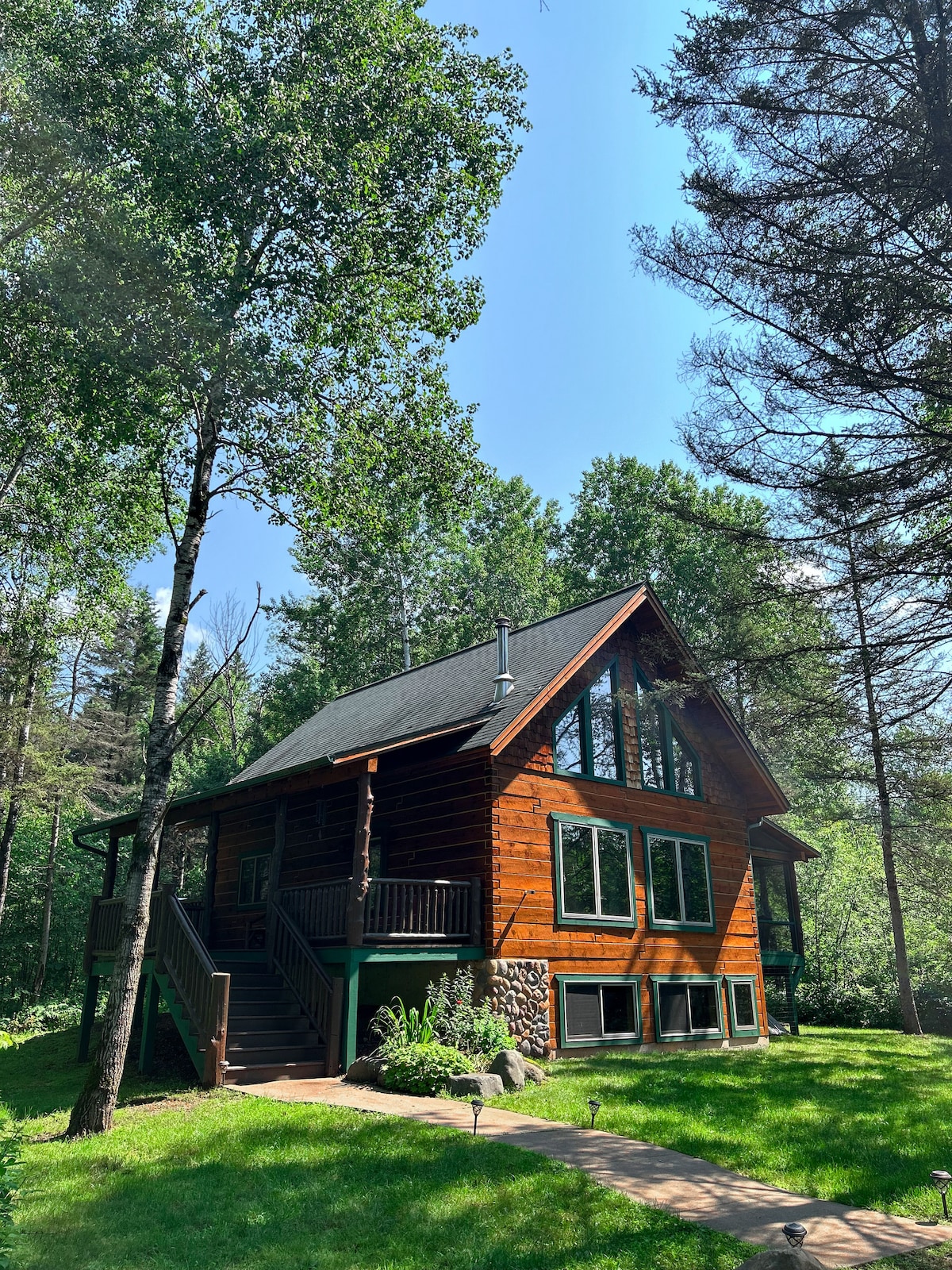 A rustic lodge is set amidst lush greenery, featuring a wooden exterior with large windows that allow natural light to fill the interior. A pathway leads up to the entrance, surrounded by well-kept landscaping and tall trees that provide a serene atmosphere.