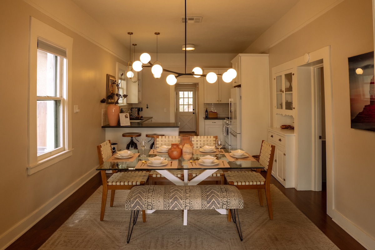 The dining area features a modern glass table surrounded by stylish wooden chairs with woven seats. A unique light fixture hangs above, and the open kitchen can be seen in the background, highlighting the contemporary design and ample natural light from nearby windows.