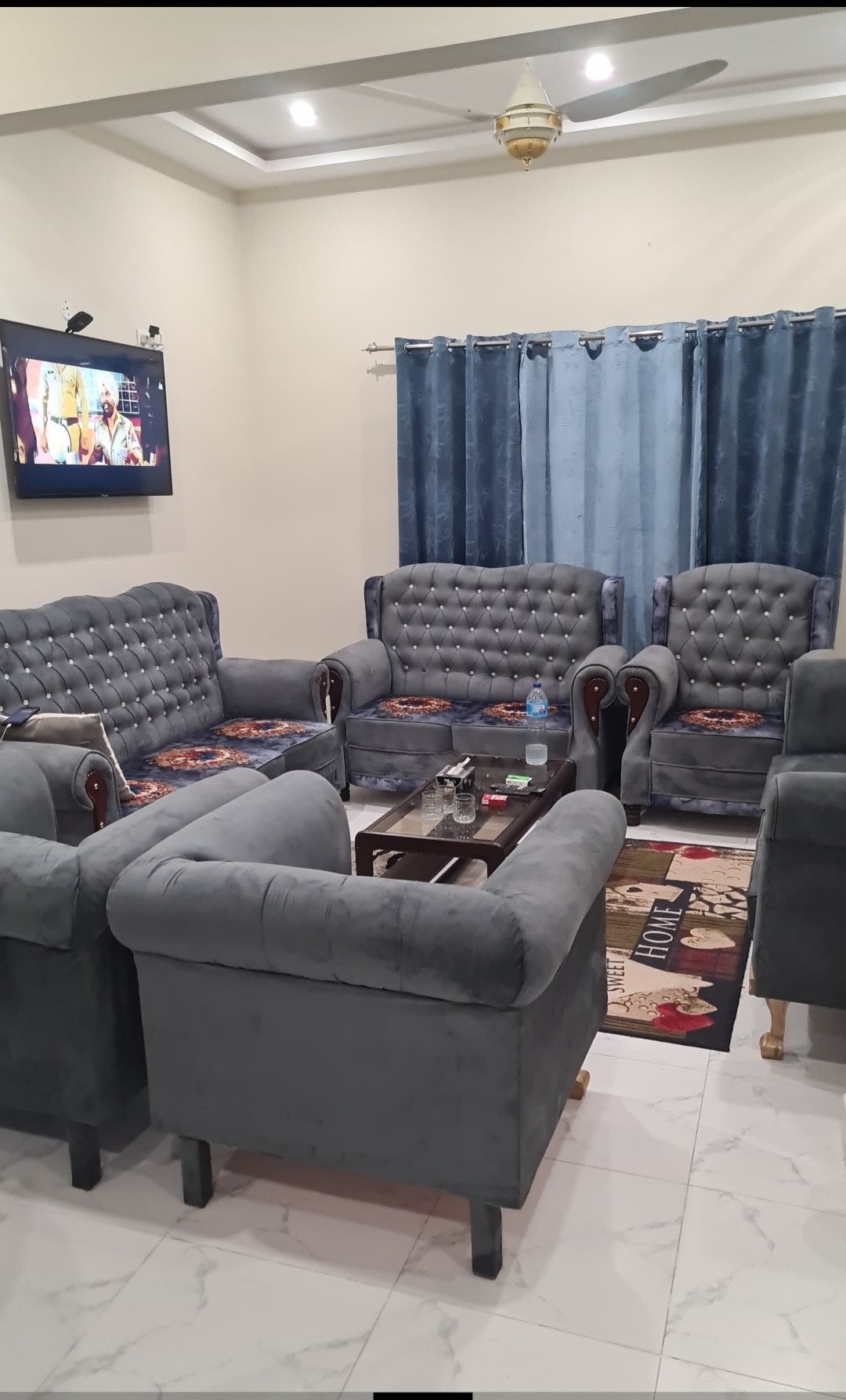 A comfortable living area features plush gray sofas arranged around a dark coffee table. A mounted television is positioned on the wall, and light curtains frame a window. A carpet with a bold pattern adds texture to the tiled floor, creating a welcoming environment.