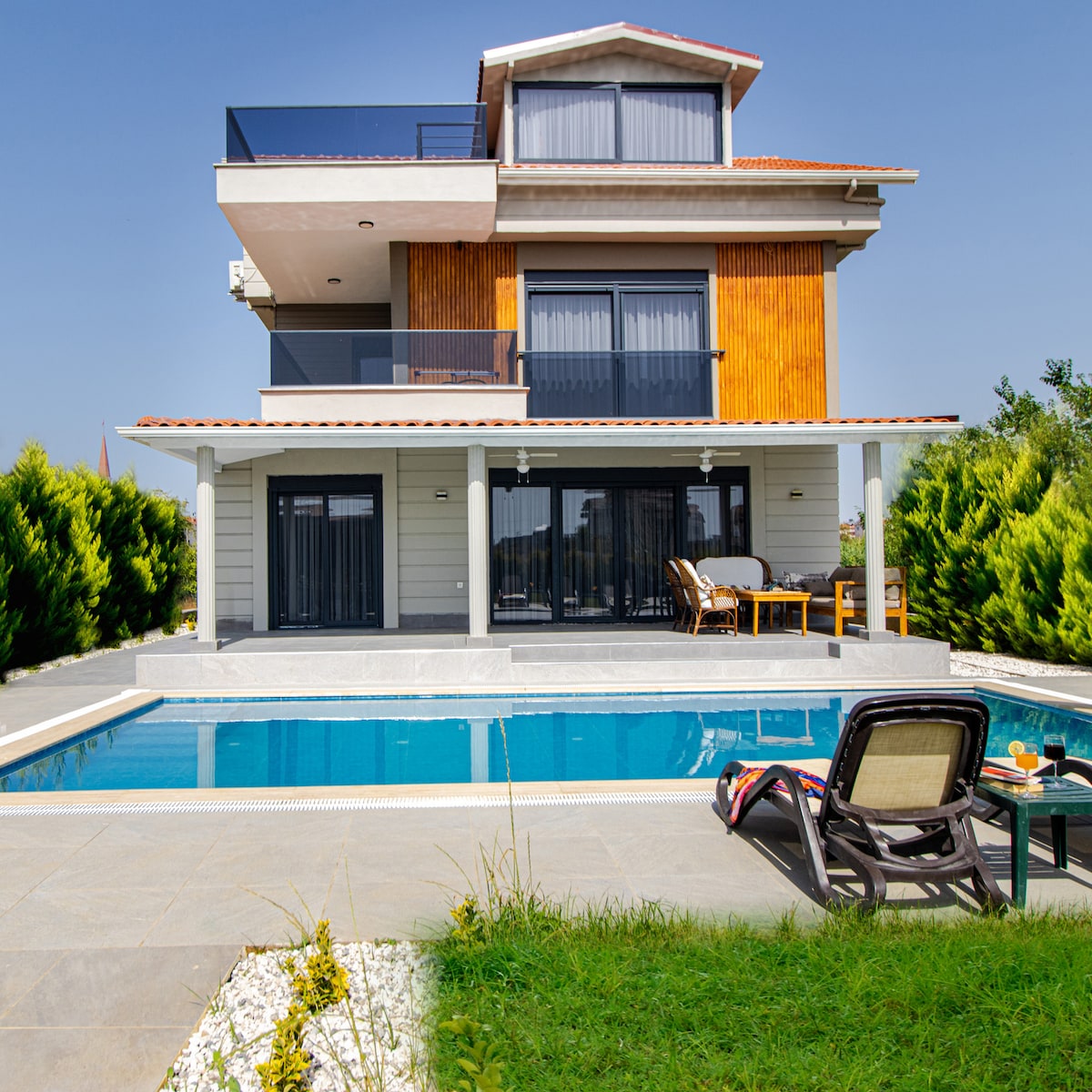The villa's contemporary exterior is highlighted by wooden accents and large glass windows. A private swimming pool reflects the clear blue sky, surrounded by sun loungers. Lush green hedges provide privacy, while an outdoor seating area offers a perfect spot for relaxation by the water.