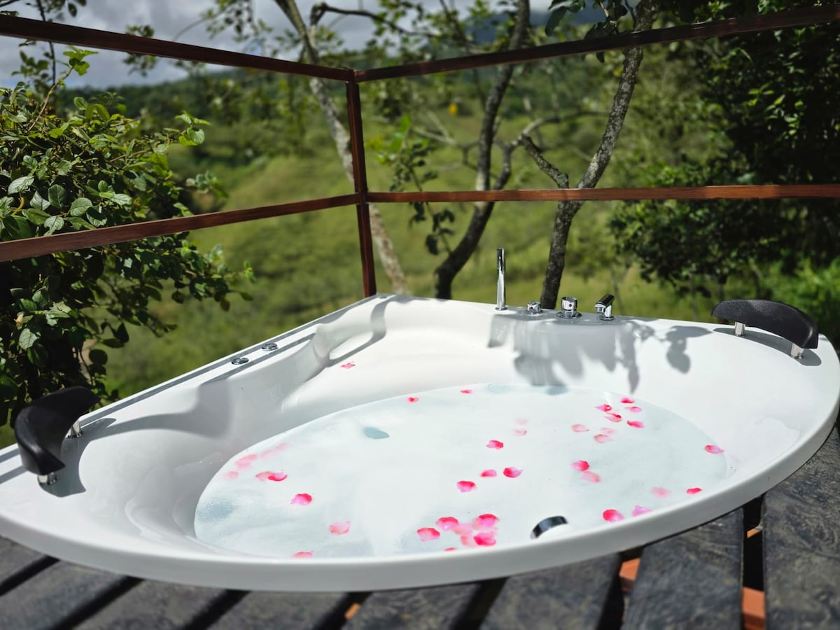 A sunken outdoor bathtub is set on a wooden deck, decorated with rose petals floating on the surface. The background features lush greenery, creating a serene atmosphere that enhances relaxation in nature.