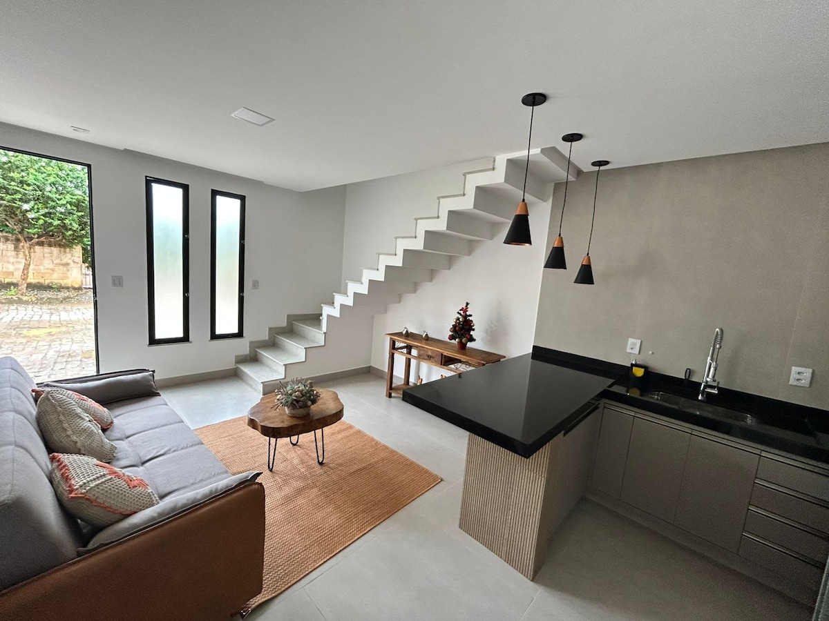 A modern living area features a comfortable sofa paired with a circular coffee table on a textured rug. The kitchen is equipped with sleek cabinets and a dark countertop. Large windows allow natural light, while a staircase leads to the upper level.