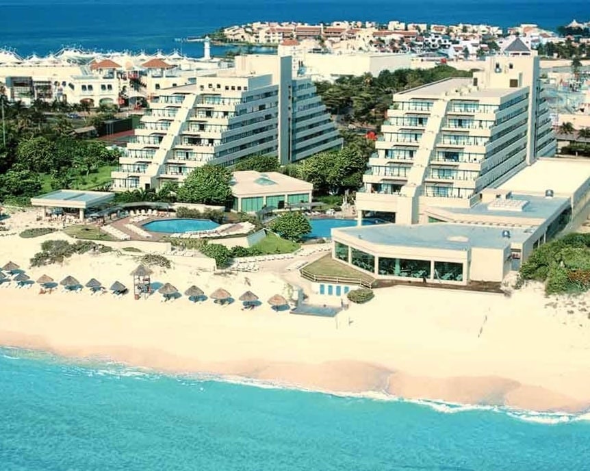 An aerial view of a beachside resort showcases multiple modern buildings arranged around inviting swimming pools and lush landscaping. The beach with soft sand and serene waves lies just steps away, with a line of umbrellas providing shaded seating along the coast.