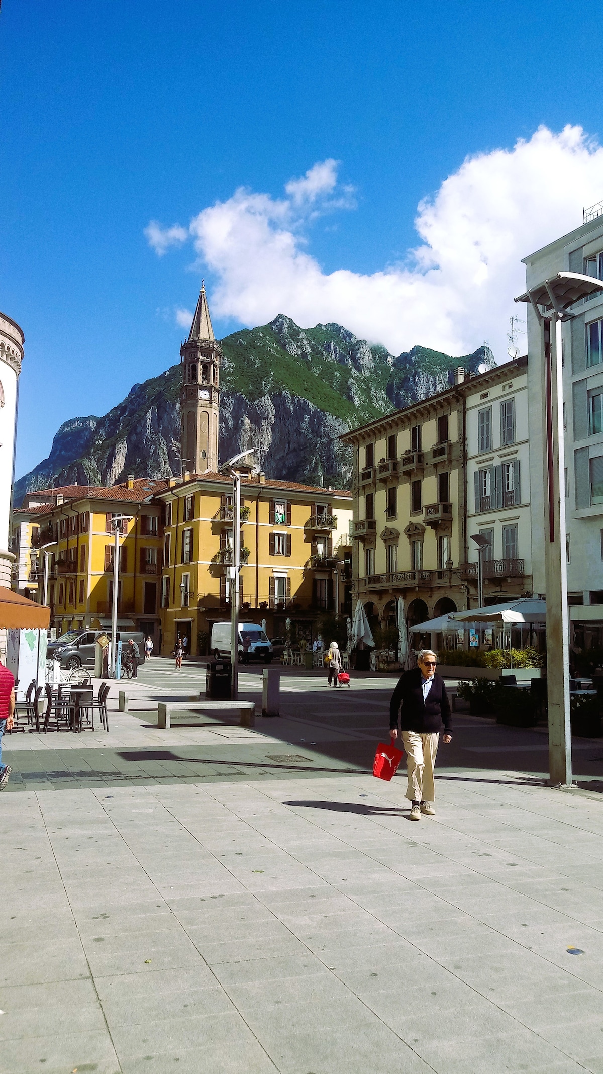 A vibrant plaza is visible, lined with colorful buildings and cafes, with outdoor seating. A church steeple rises prominently against a backdrop of green mountains. A blue sky with scattered clouds enhances the lively atmosphere of the area, often frequented by visitors and locals.