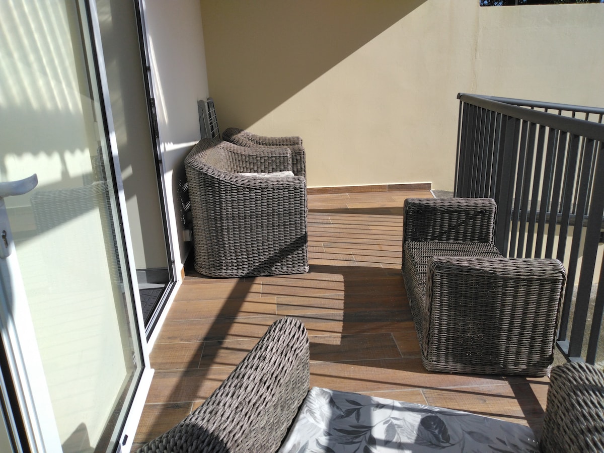 A shaded terrace space is shown, featuring a set of wicker seating arranged in a circular formation. The wooden floor adds a warm touch, while sunlight casts soft shadows across the area. Glass doors provide access to the indoors.