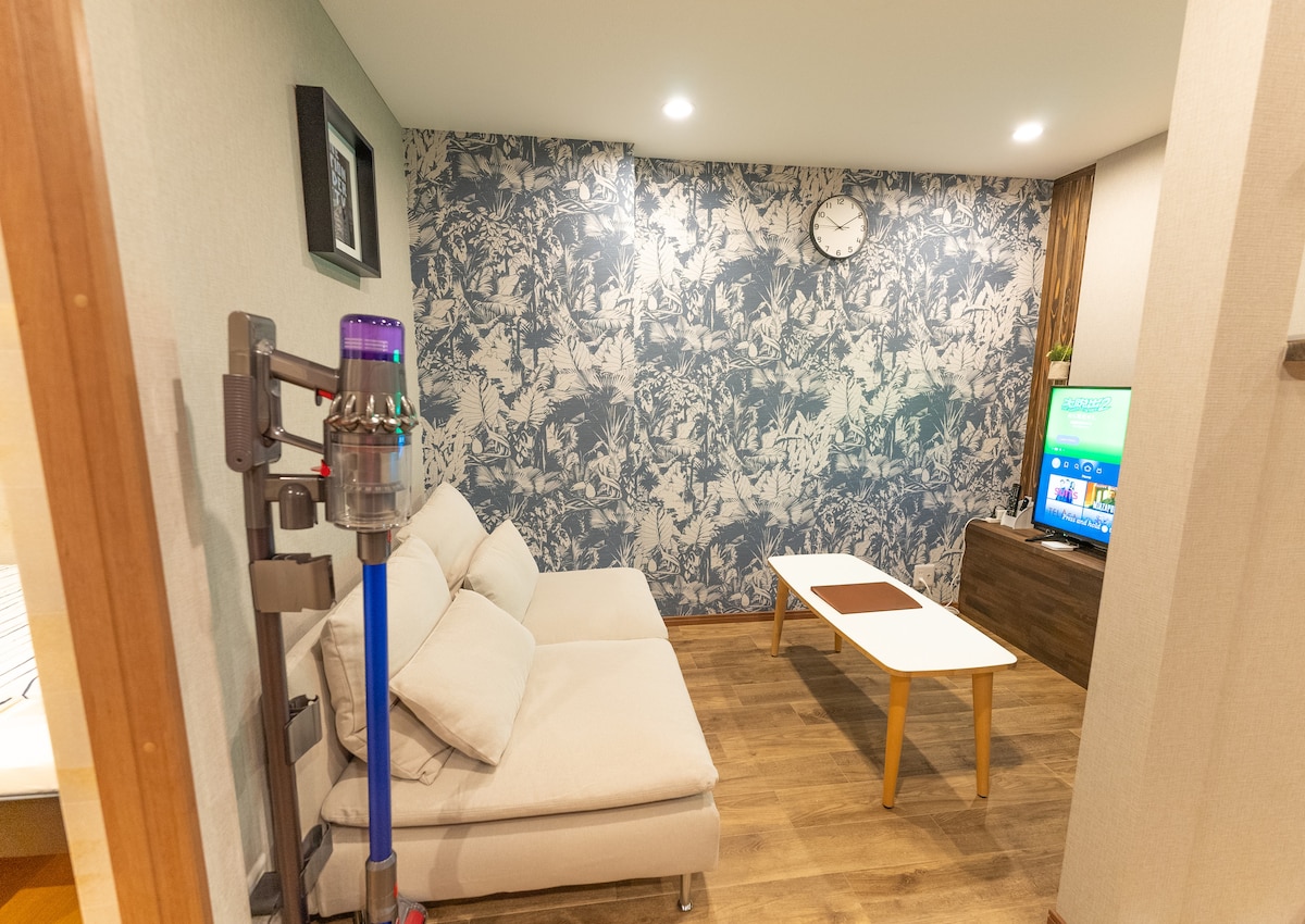 A modern living area is shown, featuring a light-colored sectional sofa positioned against a patterned wall. A clock hangs above the sofa, while a flat-screen television rests on a wooden cabinet. A small table sits in front of the seating area, and warm lighting enhances the space.