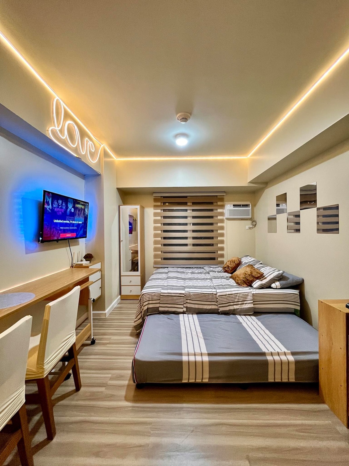 The studio features a double bed and a semi double pullout, positioned in a space with warm ceiling lighting. A Smart TV is mounted on the wall, while decorative mirrors and a desk area complete the layout. Air conditioning ensures a comfortable environment.