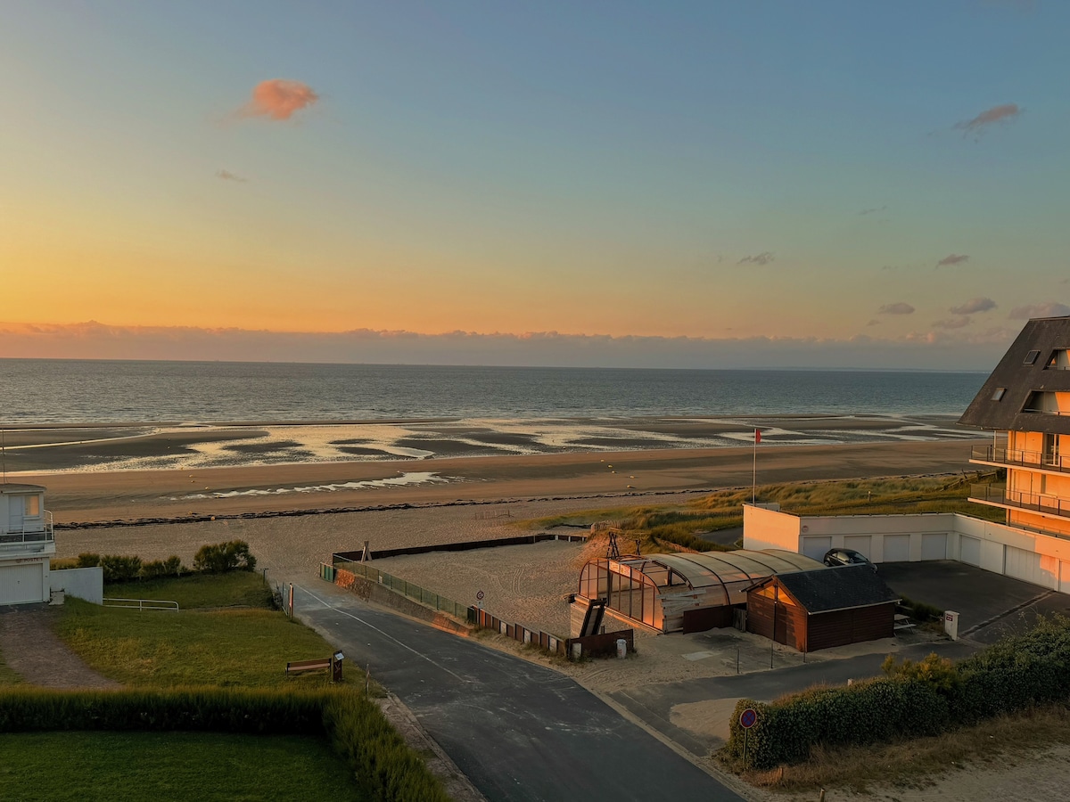 A serene view of the sandy beach at sunset, with soft waves gently lapping the shore. The colors of the sunset reflect on the horizon, creating a calming atmosphere. Nearby structures and grassy areas frame the landscape, enhancing the coastal setting.