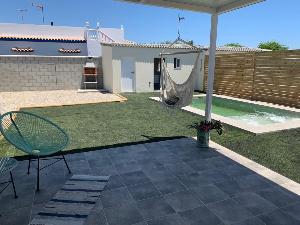 An outdoor area is surrounded by well-maintained grass and features a refreshing pool. A covered patio provides shade, with hanging seating visible. A barbecue area is positioned nearby, complemented by a light-colored stone wall and a wooden fence.