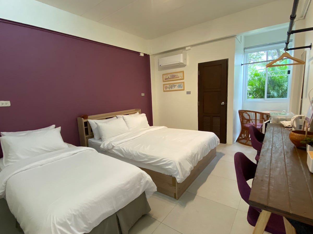 The room features two spacious beds with crisp white linens against a deep purple accent wall. A wooden work desk and purple chairs provide a functional area, while a window allows natural light to illuminate the space. Simple decorative elements enhance the room’s cozy feel.