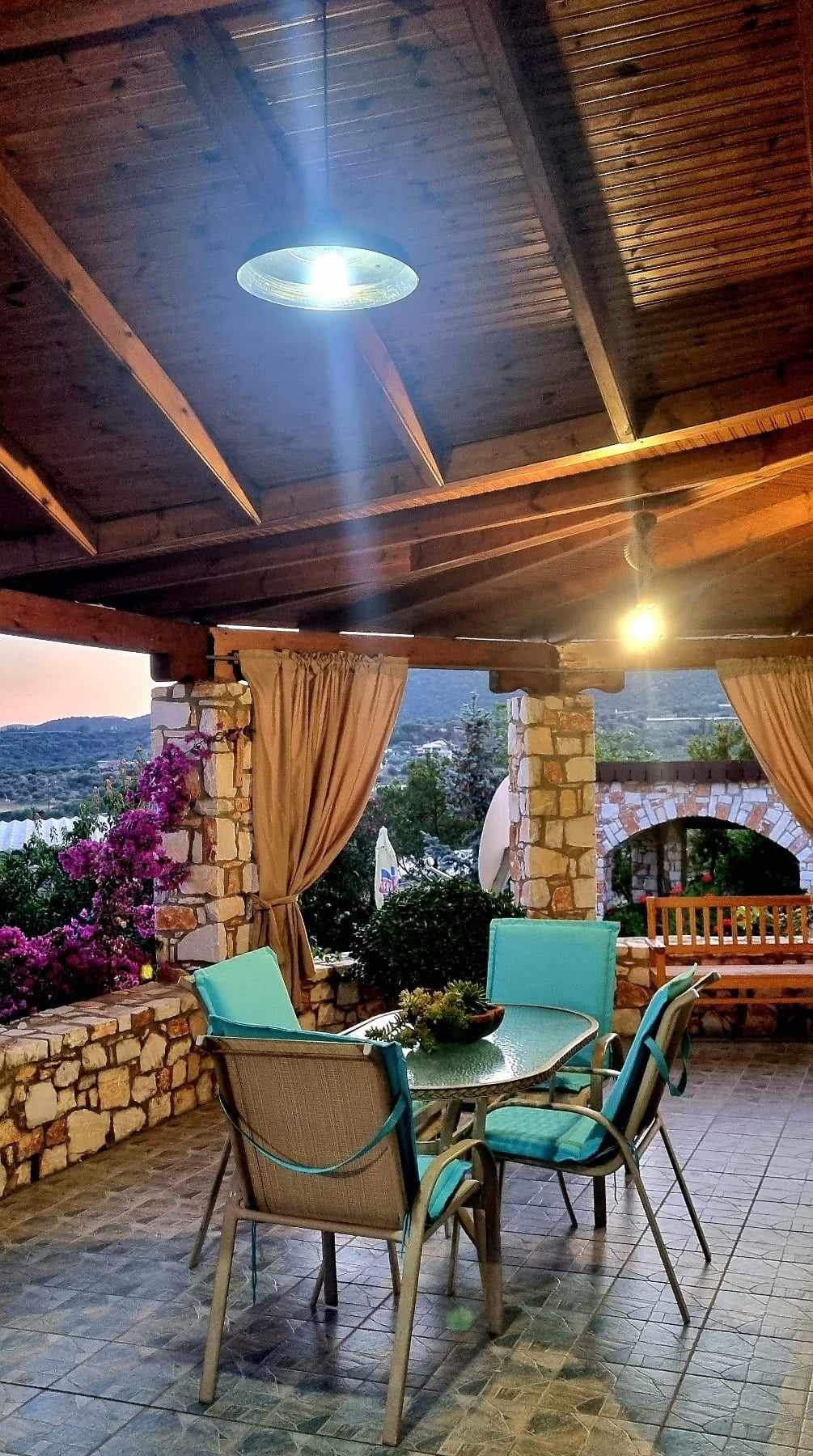 An outdoor dining area is framed by stone columns and wooden beams, illuminated by pendant lights. A round table with six chairs is surrounded by vibrant flowering plants, offering a serene space for meals overlooking the garden and sea.