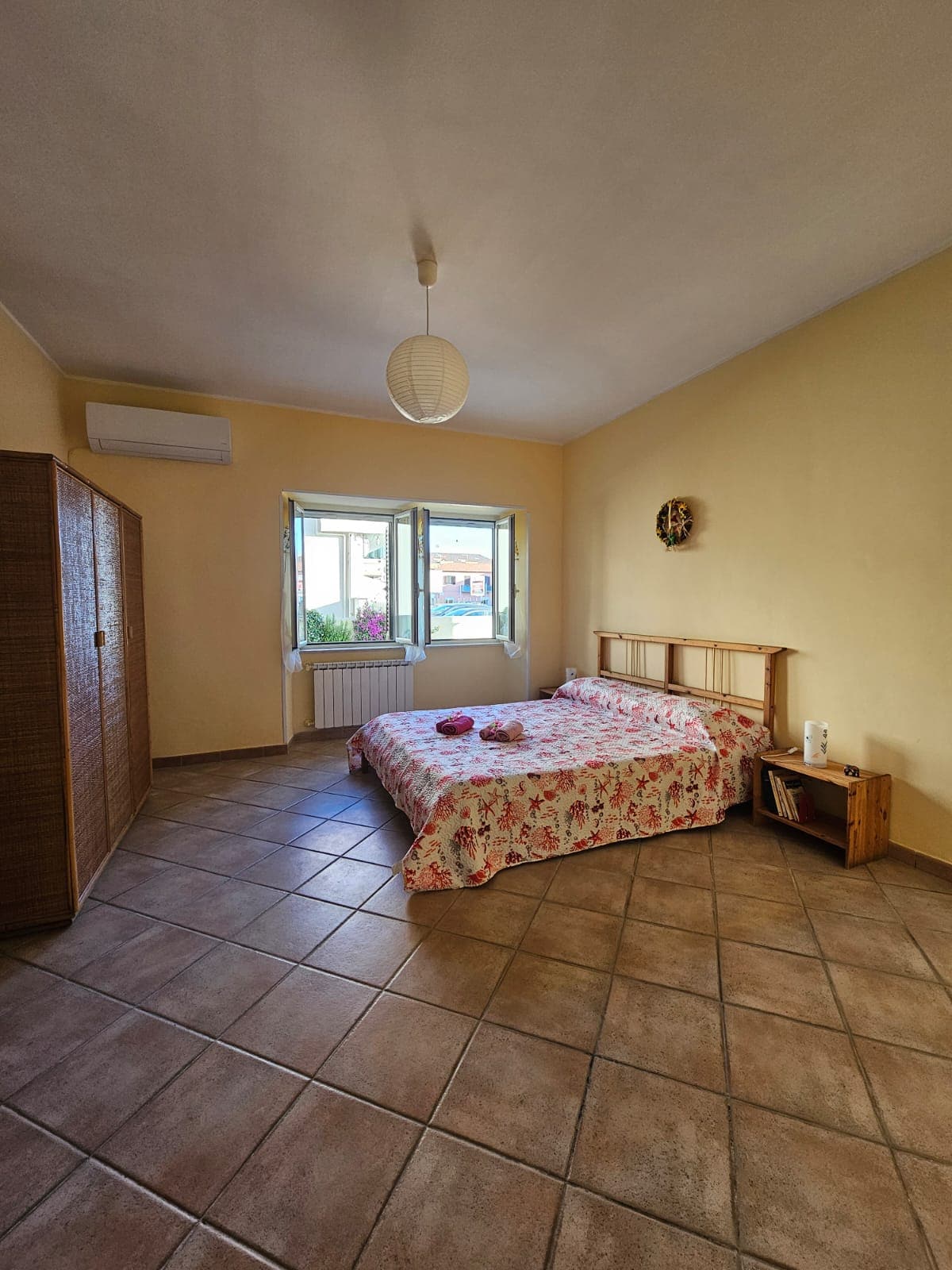 A well-lit bedroom features a spacious bed adorned with floral bedding. Large windows provide natural light and views of greenery outside. A wooden wardrobe and a small bedside table are included in the minimalist décor, complementing the neutral colored walls and tiled flooring.