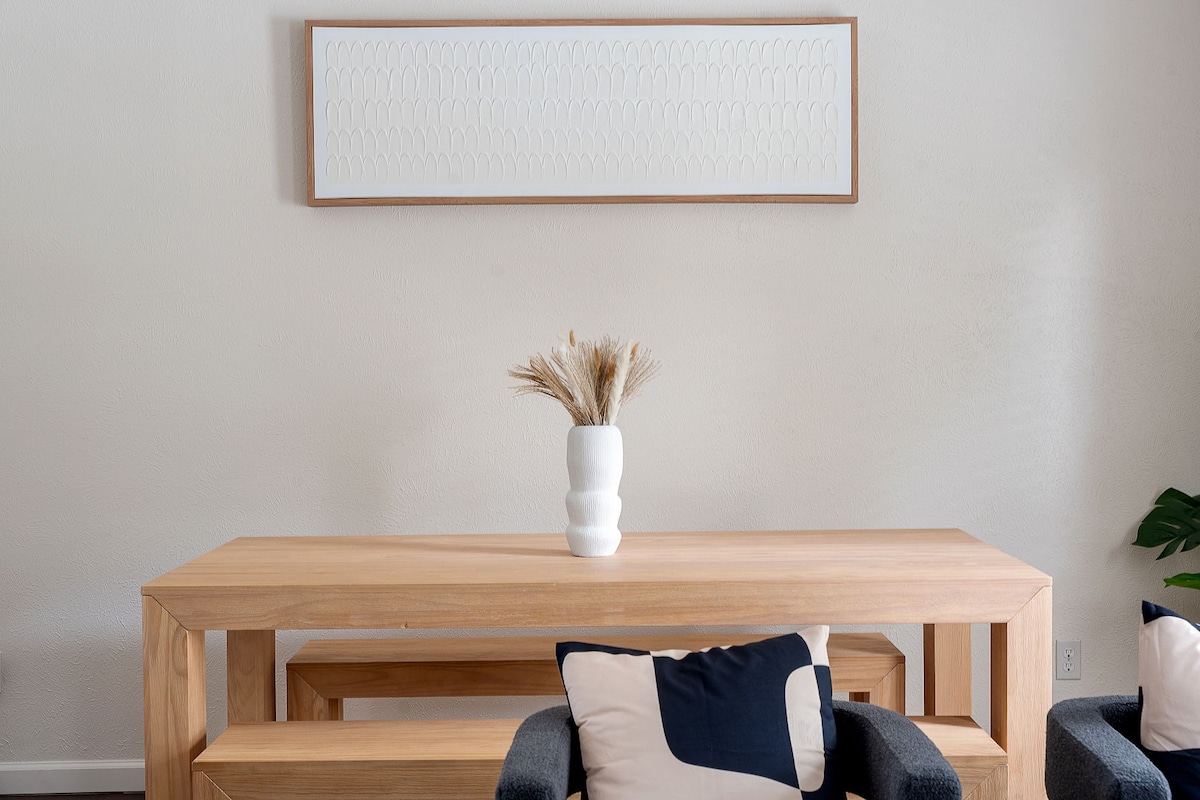 A modern wooden console table is positioned against a beige wall, showcasing a white ceramic vase filled with dried grasses. An abstract wall art piece in a light frame hangs above, complementing the neutral colors. Two chairs with dark upholstery add to the contemporary decor.