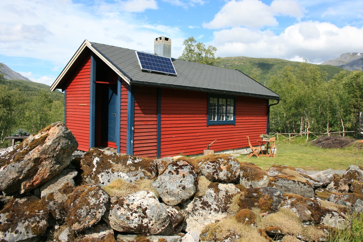 A rustic cabin with a vibrant red exterior is set against a backdrop of rolling hills and blue sky. Solar panels are mounted on the roof, while a stone wall surrounds the property. Wooden chairs are positioned outside, inviting relaxation in the natural surroundings.