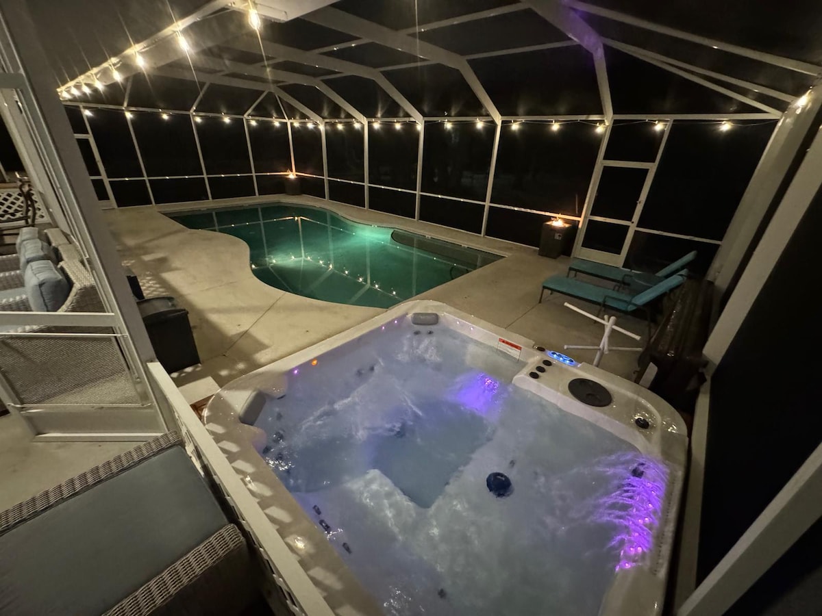 An illuminated outdoor space features a private pool and spa. Soft purple lights highlight the spa's bubbling water, while warm string lights create a serene atmosphere. The screened enclosure provides protection, ensuring a comfortable retreat for relaxation and leisure.
