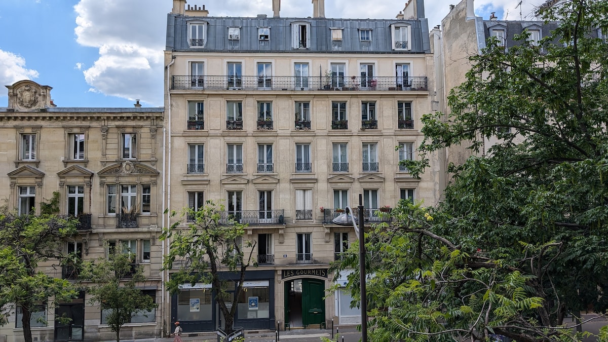 A classic Haussmannian building features a symmetrical façade with multiple balconies adorned with wrought iron railings. Lush green trees frame the view, creating a contrast against the light-colored stone exterior. The sky is filled with clouds, adding depth to the scene.