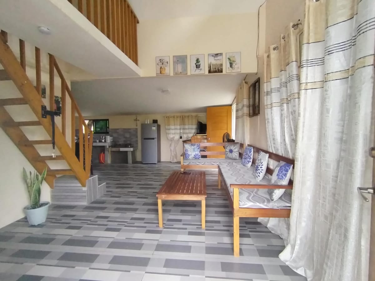 A spacious living area features wooden seating with decorative cushions, positioned beside large windows adorned with light curtains. A staircase leads to an upper level, while a sleek kitchen appliance is visible in the background, complementing the modern decor.