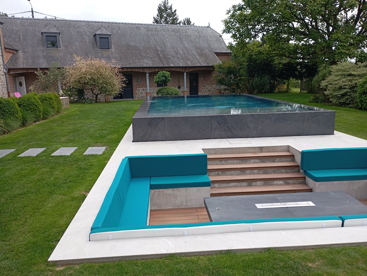 A modern infinity pool is set within a manicured lawn, featuring a built-in seating area with turquoise cushions and wooden steps leading into the water. The rustic stone house is visible in the background, surrounded by lush greenery.