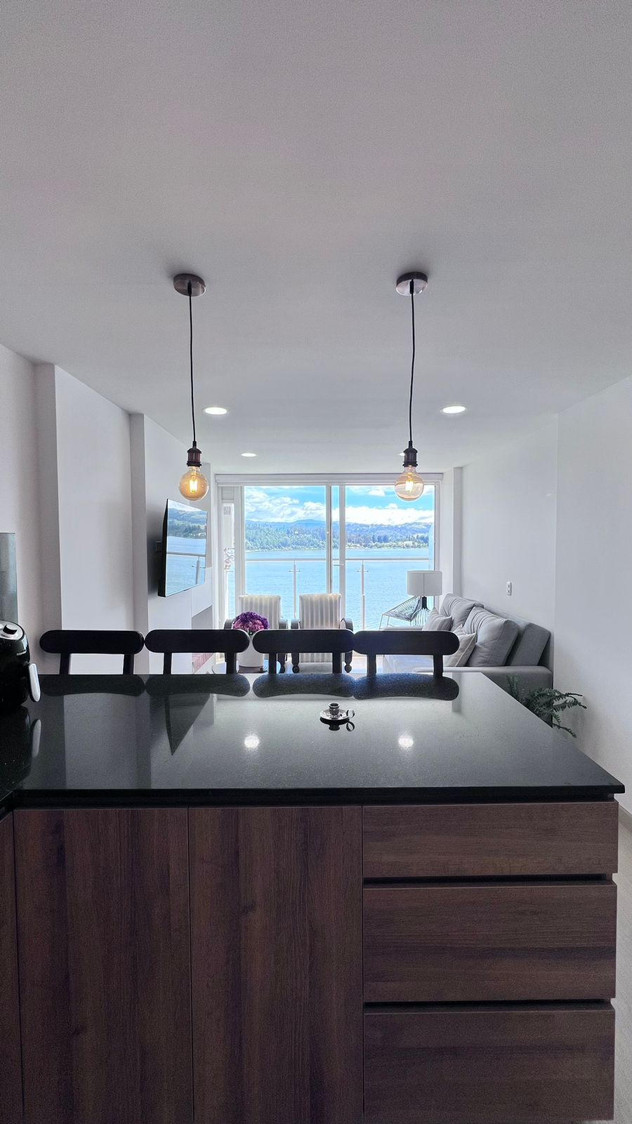 A modern kitchen area features a dark countertop with minimalist wooden cabinetry. Two pendant lights hang above, providing soft illumination. In the background, a spacious living area opens to large glass windows revealing scenic views of the lake, complemented by natural light streaming in.
