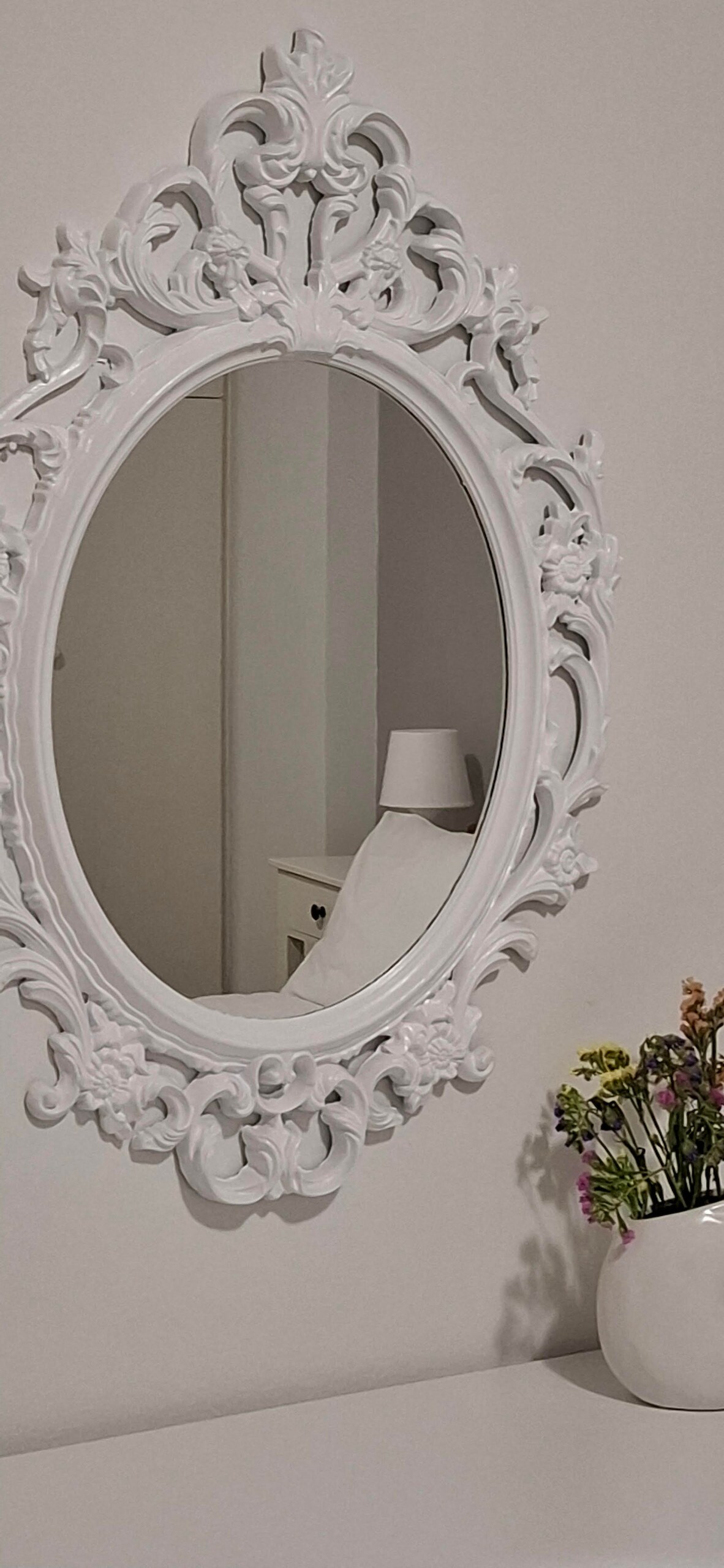 An ornate white mirror with intricate detailing is mounted on a neutral wall. A simple white vase holds a small arrangement of colorful flowers, complementing the decor. Soft lighting enhances the serene ambiance, suggesting a peaceful living space.