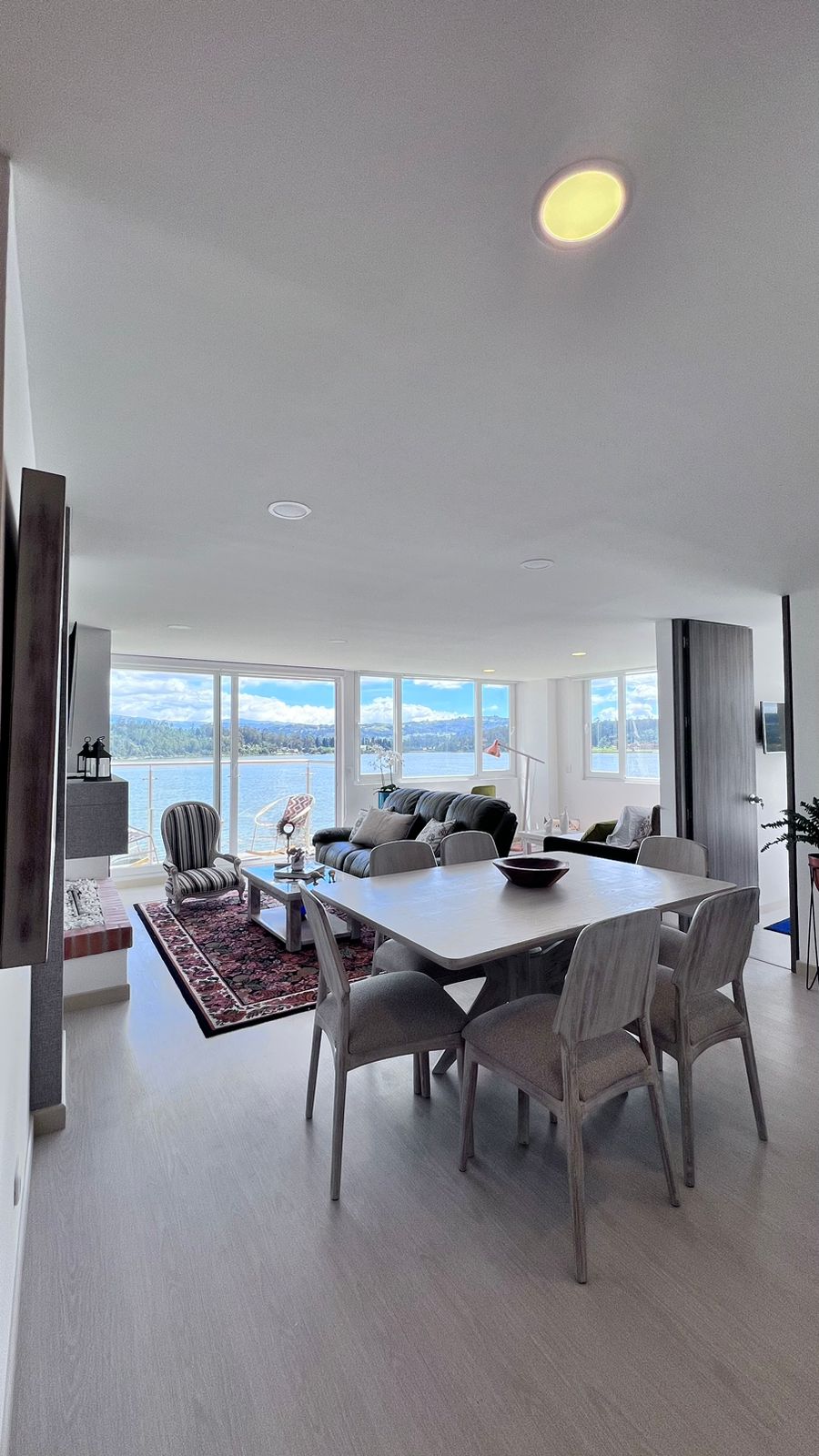 A modern living area is presented, featuring large windows that offer views of the lake and mountains. A comfortable sofa and a dark wood coffee table are arranged on a patterned area rug. A dining table with light-colored chairs complements the space, enhancing the inviting atmosphere.