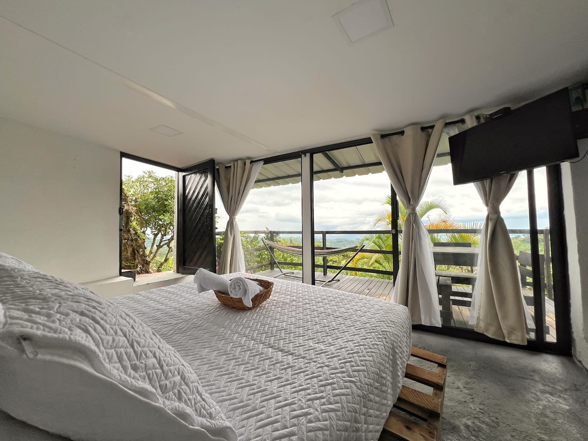A comfortable sleeping area features a spacious bed dressed in a textured white coverlet. Large glass sliding doors open to a private balcony, enhancing natural light and providing views of surrounding landscapes. Soft curtains frame the windows, creating a serene atmosphere.