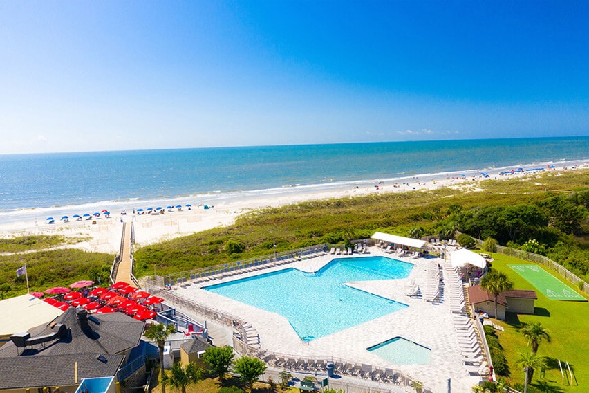 An expansive, rectangular beachfront pool is seen from above, surrounded by lounge chairs and umbrellas. The ocean stretches in the background under a clear blue sky, with a sandy beach leading to the water. Pathways are visible, providing access to the beach.