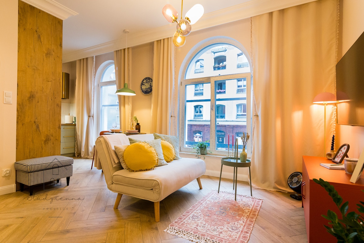 A cozy living space is highlighted by large arched windows that allow natural light to flow in. A light-colored sofa, accented with colorful cushions, is positioned near a small table. Wooden accents and soft curtains contribute to a warm atmosphere.