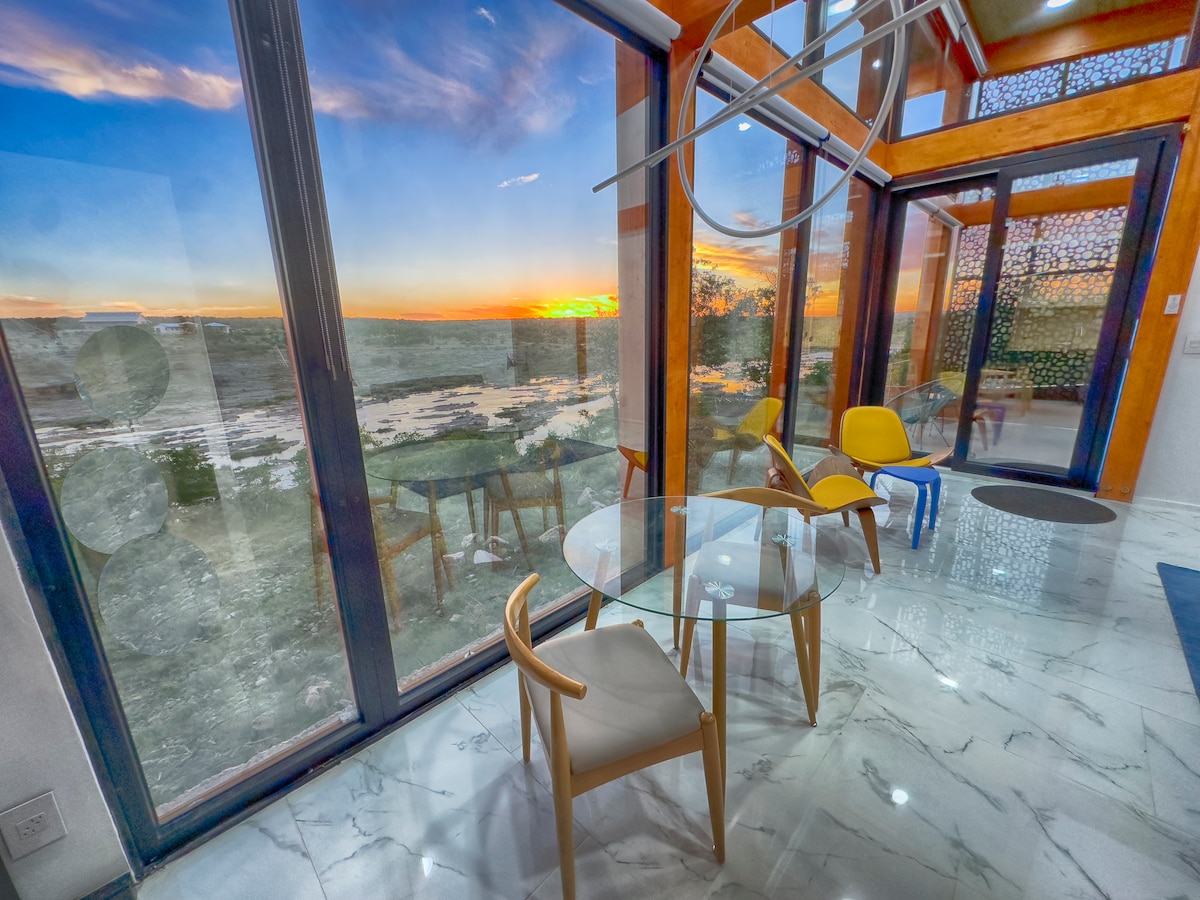 A dining area features a glass table surrounded by four wooden chairs, with a scenic view visible through large glass windows. The colors of the sunset provide a vibrant backdrop, enhancing the serene atmosphere of the space.
