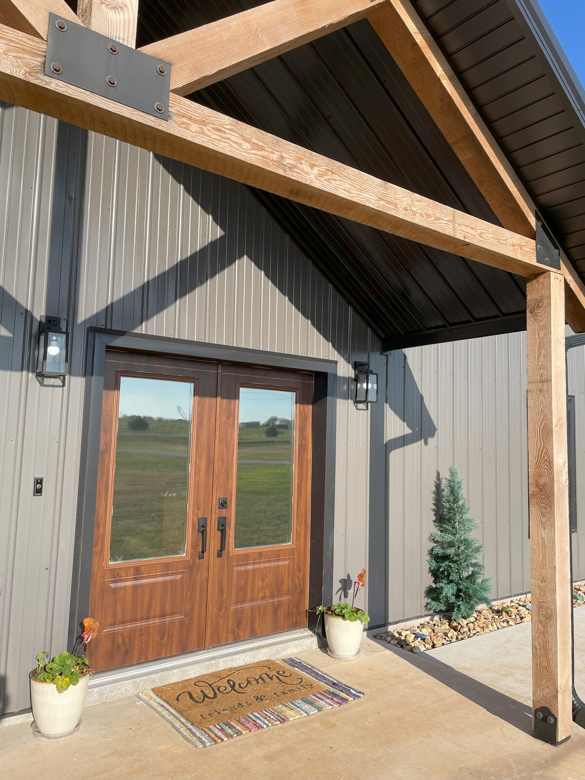 The entryway features a large wooden double door framed by two stylish wall sconces, casting soft light. A welcoming doormat displays the word 'Welcome.' Potted plants and river stones add natural elements to the modern exterior, complemented by a glimpse of green landscaping beyond.