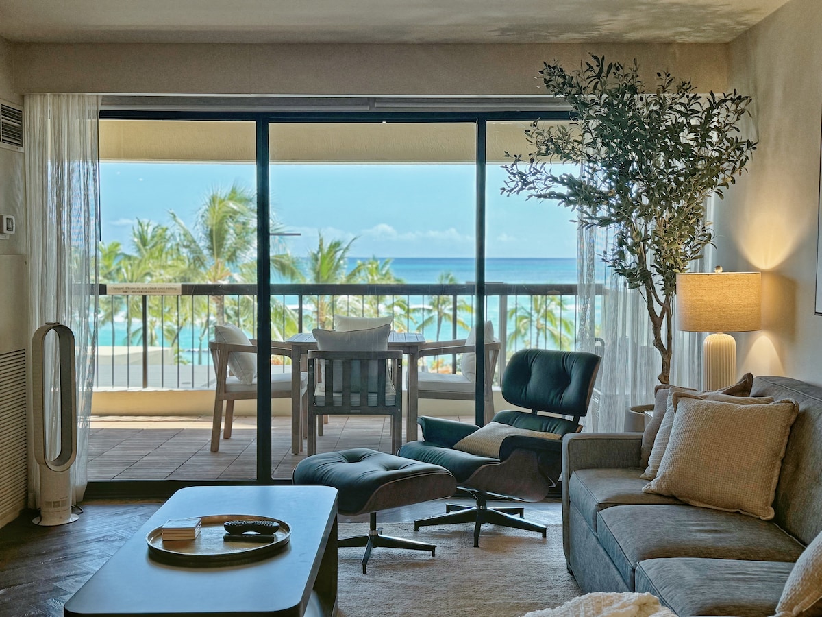 A comfortable living area features a spacious sofa and a stylish lounge chair, facing large glass doors that open to a private balcony. The ocean and palm trees can be seen beyond the balcony, with natural light illuminating the space.