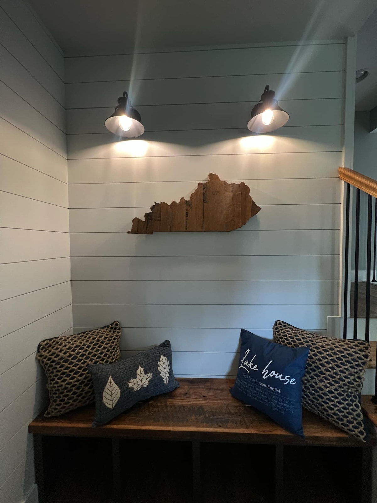 A cozy nook features a wooden bench adorned with patterned cushions in shades of blue and brown. Above, a wooden outline of Kentucky is highlighted by mounted lights, adding a subtle decorative touch to the space. Natural light enters from nearby areas.