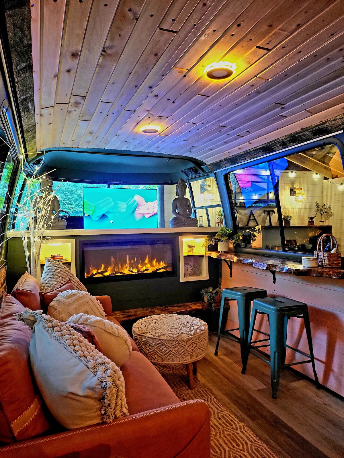 The cozy interior of the camper features a plush orange sofa adorned with decorative pillows. A sleek electric fireplace is positioned below a large television, while a compact kitchen area with bar stools is visible in the background, complemented by warm lighting and wooden accents.